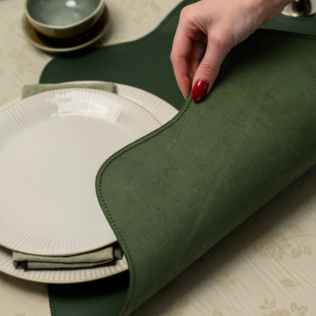 A close-up shot of a hand lifting the edge of a forest green leather placemat to reveal its soft, suede-like underside. This action highlights the thickness and premium quality of the material. The placemat features a scalloped edge with precise tonal stitching. In the background, white ribbed plates and a small bowl rest on a cream floral tablecloth, emphasizing the placemat's functional and aesthetic appeal in a table setting