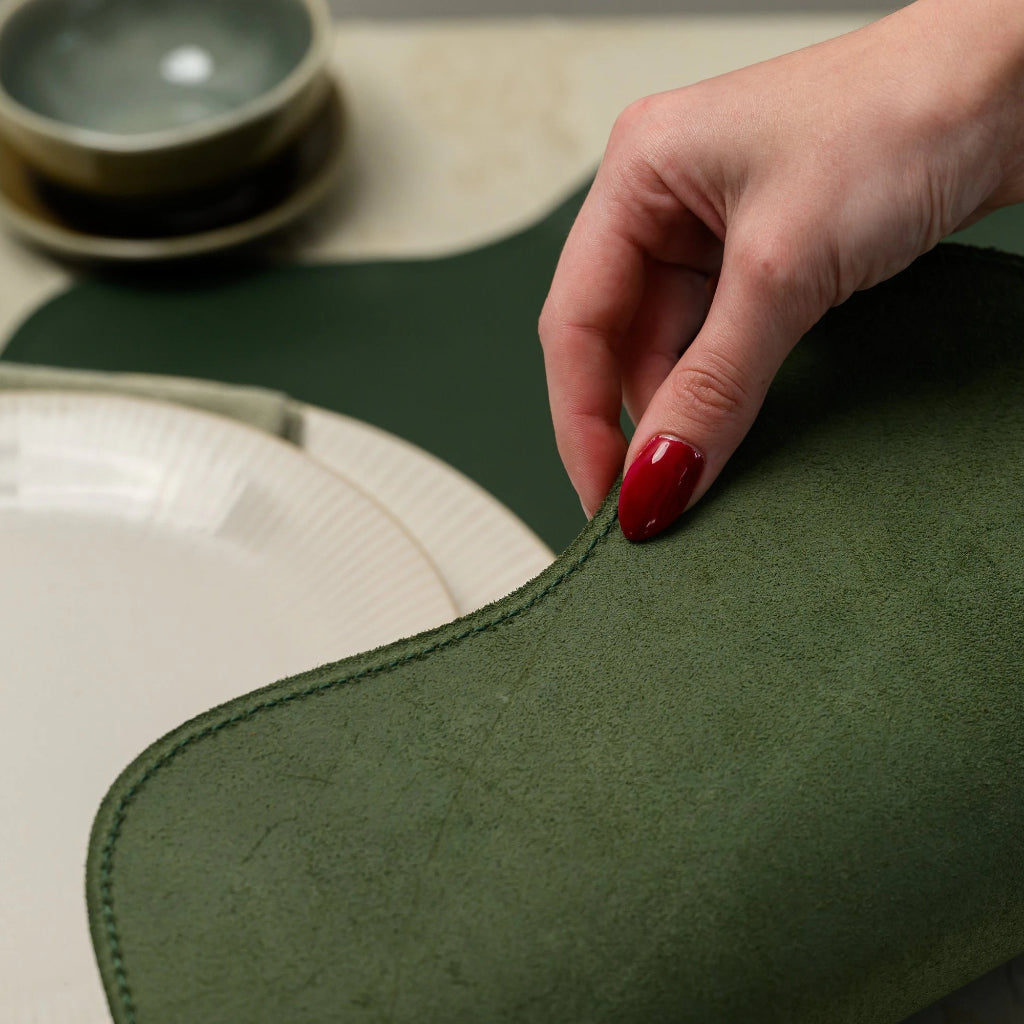 An extreme close-up focusing on the tactile quality of a dark green leather placemat. A hand with a red manicure holds the curved, scalloped edge, showcasing the soft, fibrous texture of the green suede underside and the precise perimeter stitching. The smooth top surface of the placemat and a white ribbed plate are visible in the soft-focus background, emphasizing the premium material and craftsmanship