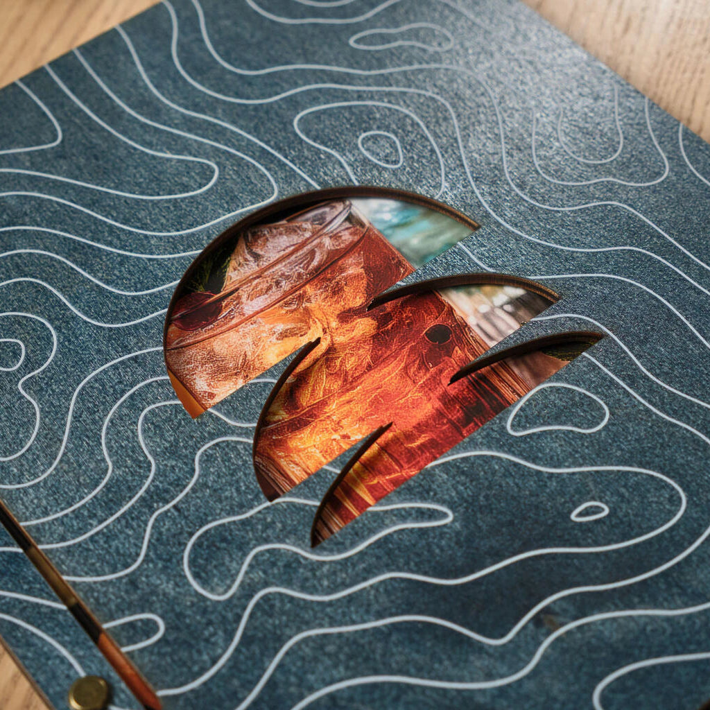 Macro close-up of a blue wooden menu cover featuring a white topographic map pattern and a precision-cut window showcasing a cocktail image