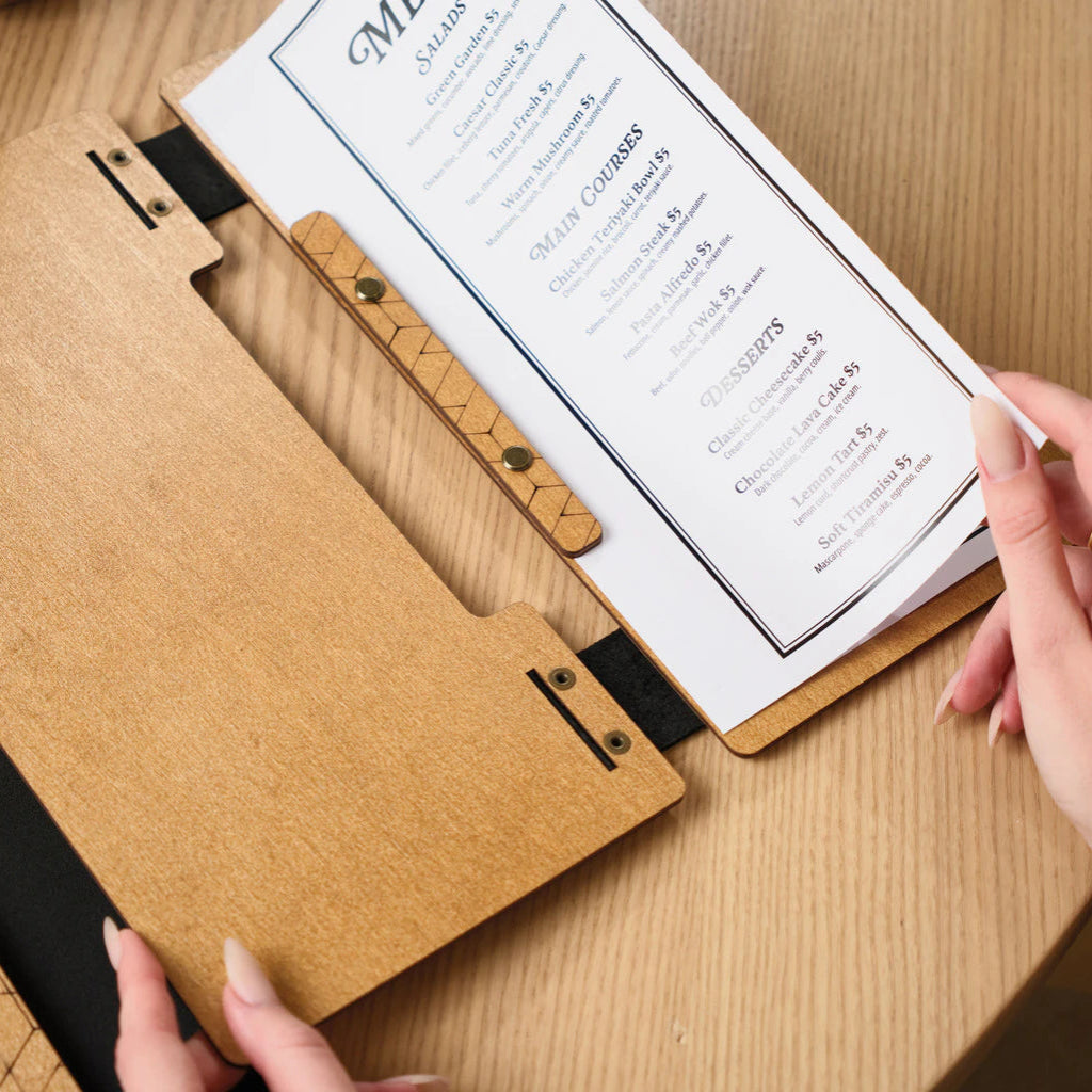 A close-up shot of hands turning the pages of a printed menu inside a wooden folder, emphasizing the durable construction and the elegant typography of the menu categories.