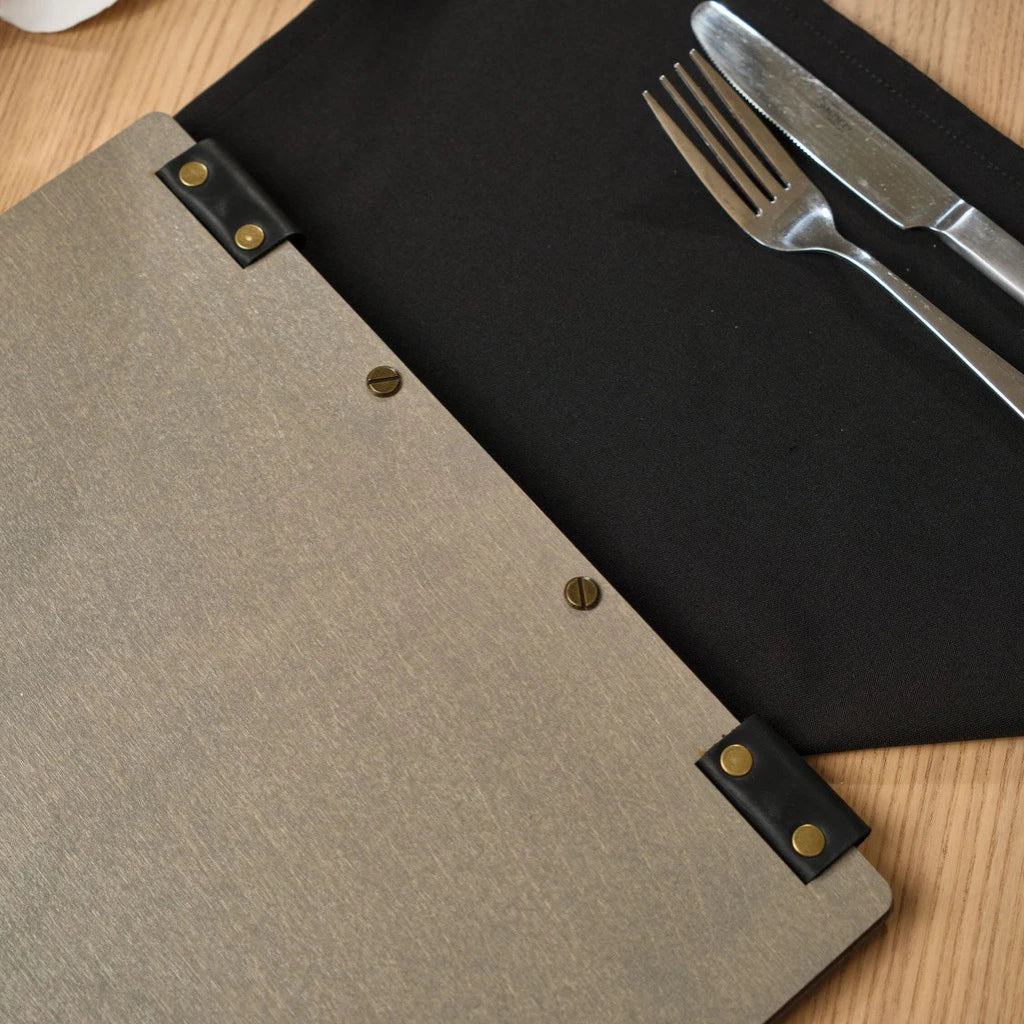 Detailed macro shot of a premium grey wood menu holder with flexible black leather hinges secured by gold-toned rivets. The image showcases the high-quality craftsmanship and durable bolt binding system, presented on a wooden table with elegant cutlery