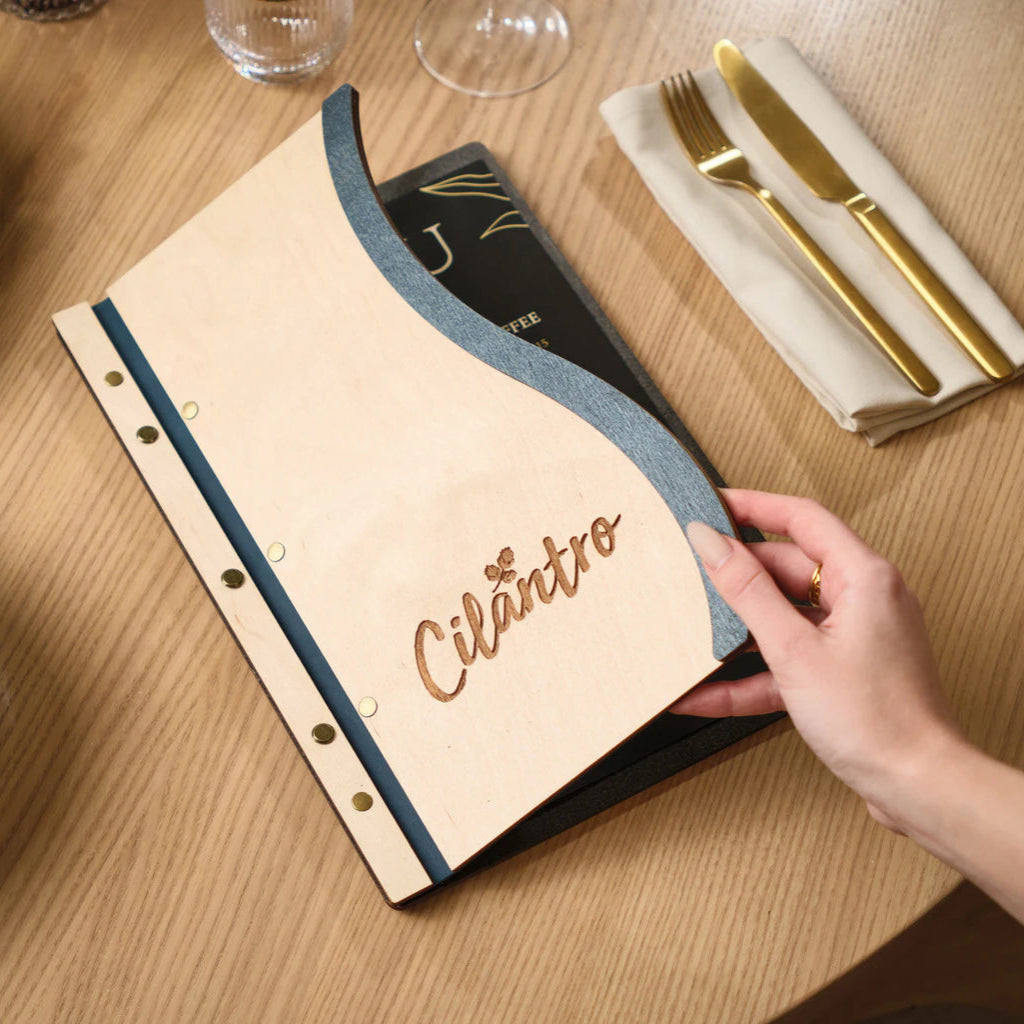 High-angle view of a hand interacting with a premium Ivory and Jeans blue wood menu folder, styled next to gold cutlery and fine glassware