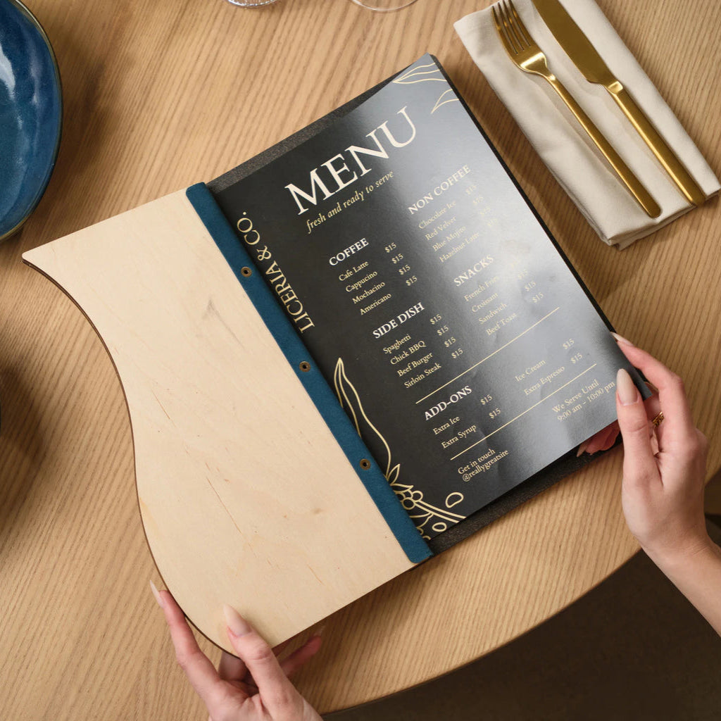 Overhead view of an open custom menu board with an Ivory wood interior and a blue Jeans-textured spine, holding a printed black menu card on a wooden table
