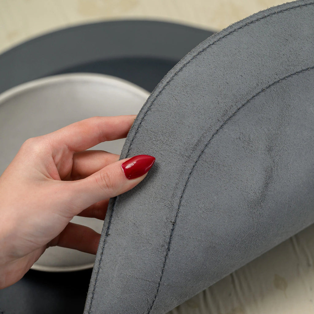 A close-up view of a hand lifting the curved edge of a leather placemat, showcasing the raw, fibrous texture of its underside. The image emphasizes the craftsmanship, specifically the visible lines of perimeter stitching that secure the layered material. A ceramic plate and the smooth top surface of the placemat are visible in the soft-focus background on a floral tablecloth