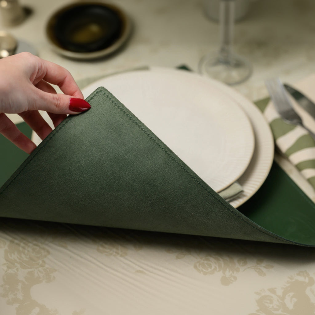 A hand lifting the corner of a square forest green leather placemat to reveal the soft suede-like backing, demonstrating the mat's flexibility and premium double-sided texture
