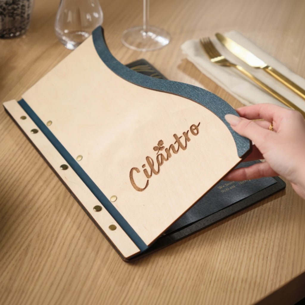 Lifestyle shot of a person opening a wavy-edged wooden menu at a table, highlighting the Ivory wood grain and the "Cilantro" engraved logo