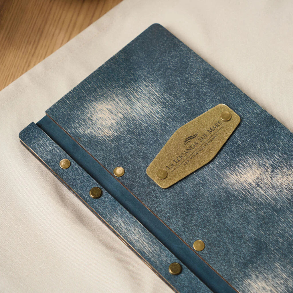 Close-up of a blue wooden menu cover featuring a brass plate with "La Locanda Sul Mare" logo