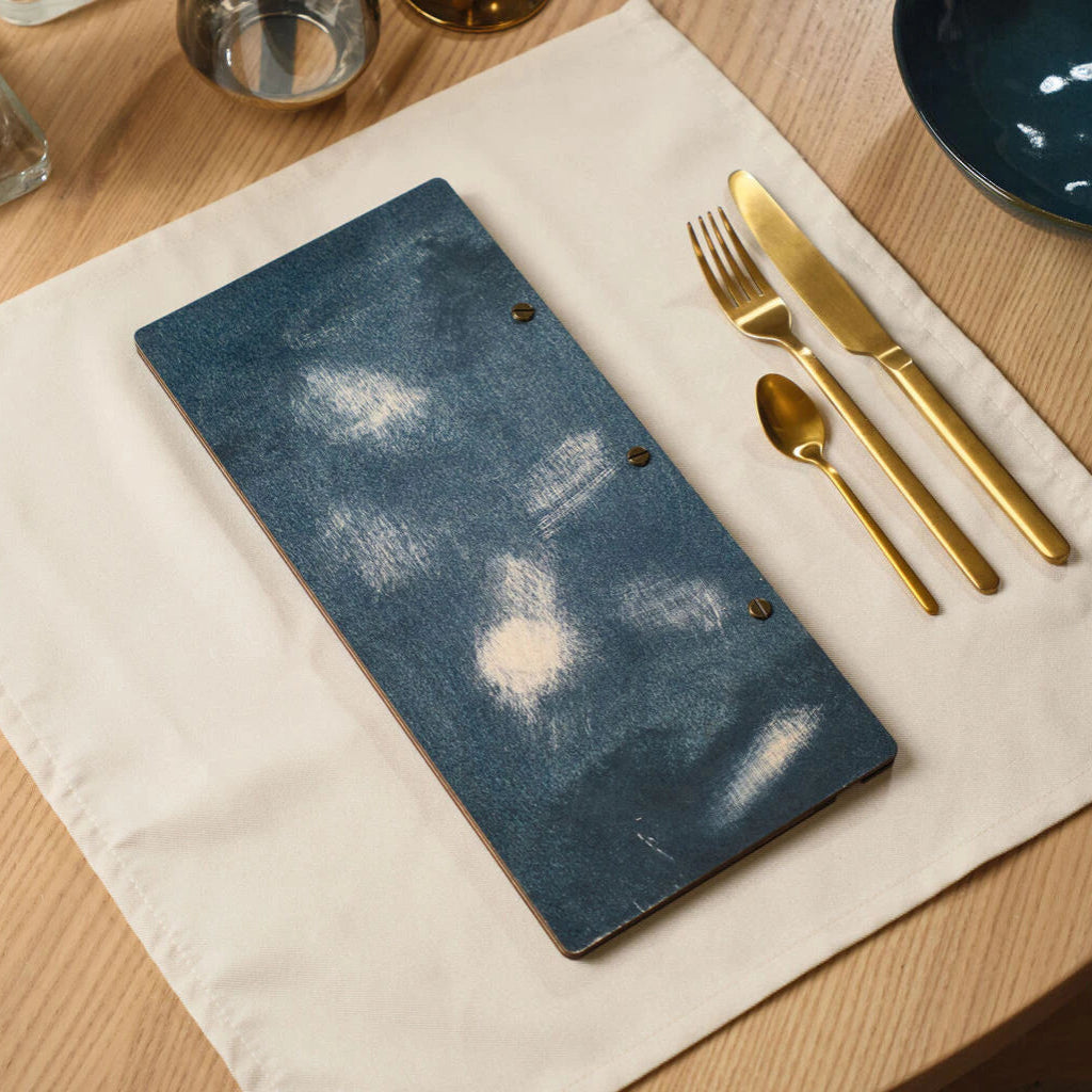 A top-down view of the slim, blue distressed wood menu folder lying on a white linen napkin next to a set of gold cutlery on a wooden restaurant table.