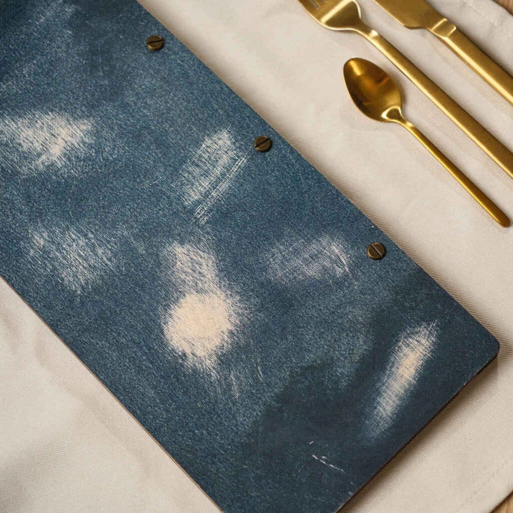 A close-up shot of a wooden menu cover in a "jeans" blue finish, featuring a distressed, weathered texture with light wood grain showing through, accented by dark slotted brass screws.