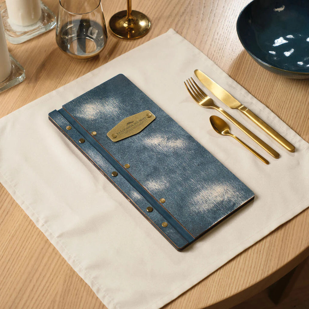 A stylish blue-tinted wooden menu holder with brass accents, placed on a table setting alongside elegant gold forks and knives.