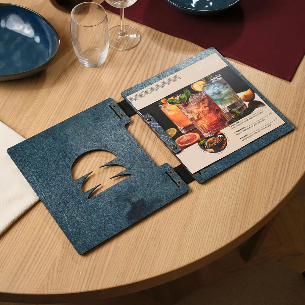 A  opening a blue wooden menu cover with a topographic map pattern, revealing the internal pages and black leather binding on a wooden restaurant table