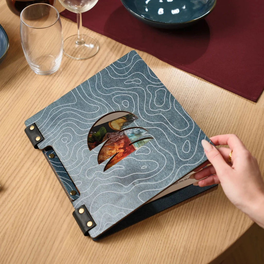 Handheld view of a custom blue menu folder featuring a unique laser-cut window and topographic line design, styled with dining glassware