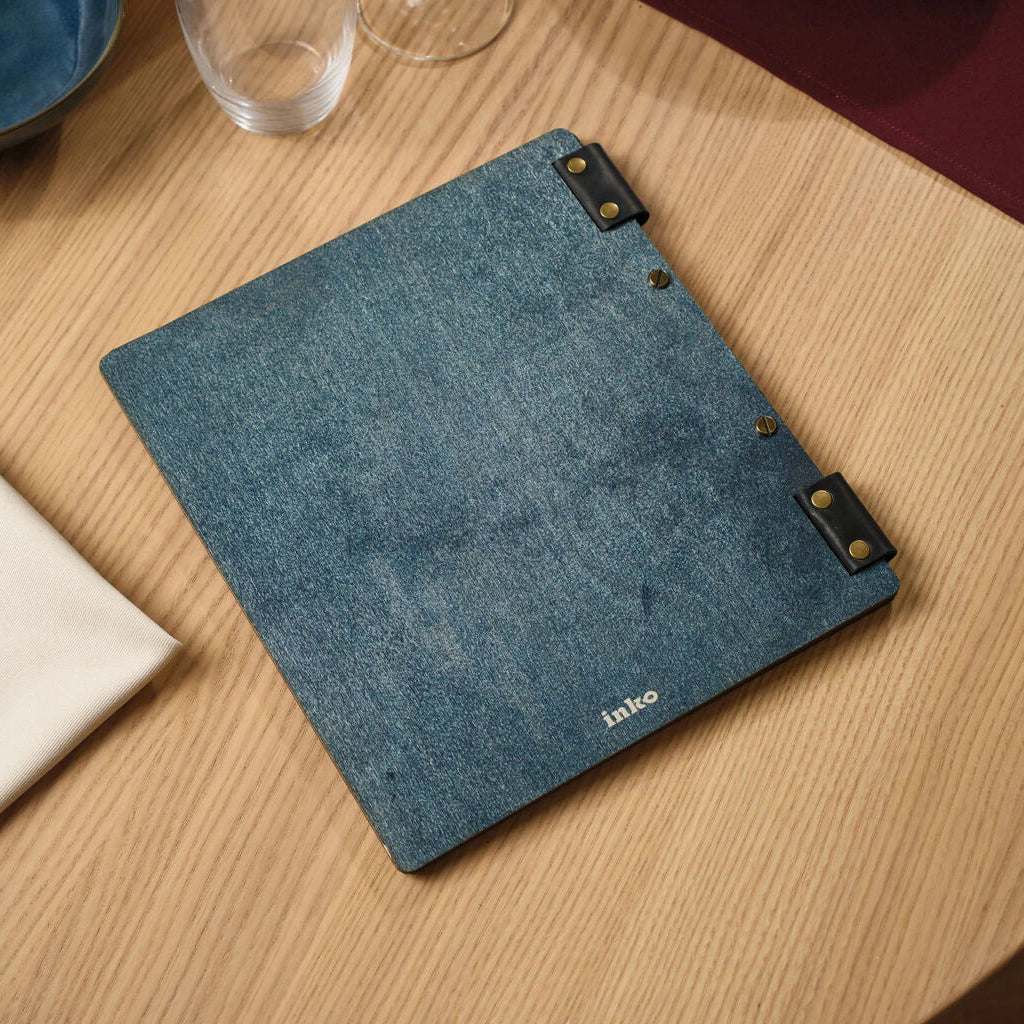 Rear view of a premium blue textured wooden menu cover, featuring a silver "ink" brand logo and two black leather straps with brass rivets on the edge