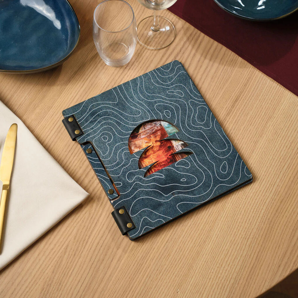 Unique topographic pattern menu holder in blue wood and leather, placed on a light oak wooden table with dining accessories