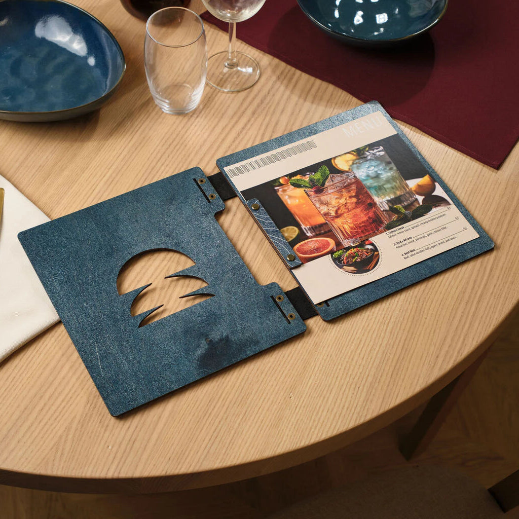 Open blue wooden menu folder with a decorative cutout on the cover, showing an internal menu page with cocktail photos secured by brass screws