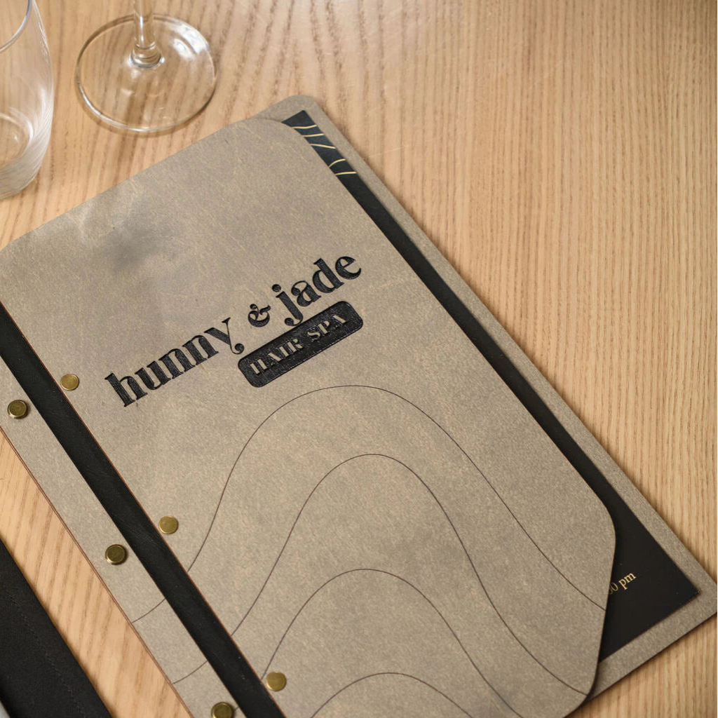 Close-up of a high-quality laser-engraved "Hunny & Jade Hair Spa" logo on a custom wooden menu cover with elegant brass hardware details