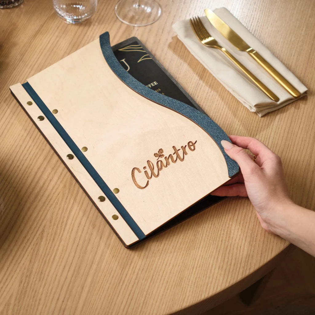 A hand lifting the front cover of a custom Ivory wood menu board, revealing a black menu insert held by a blue Jeans-textured spine