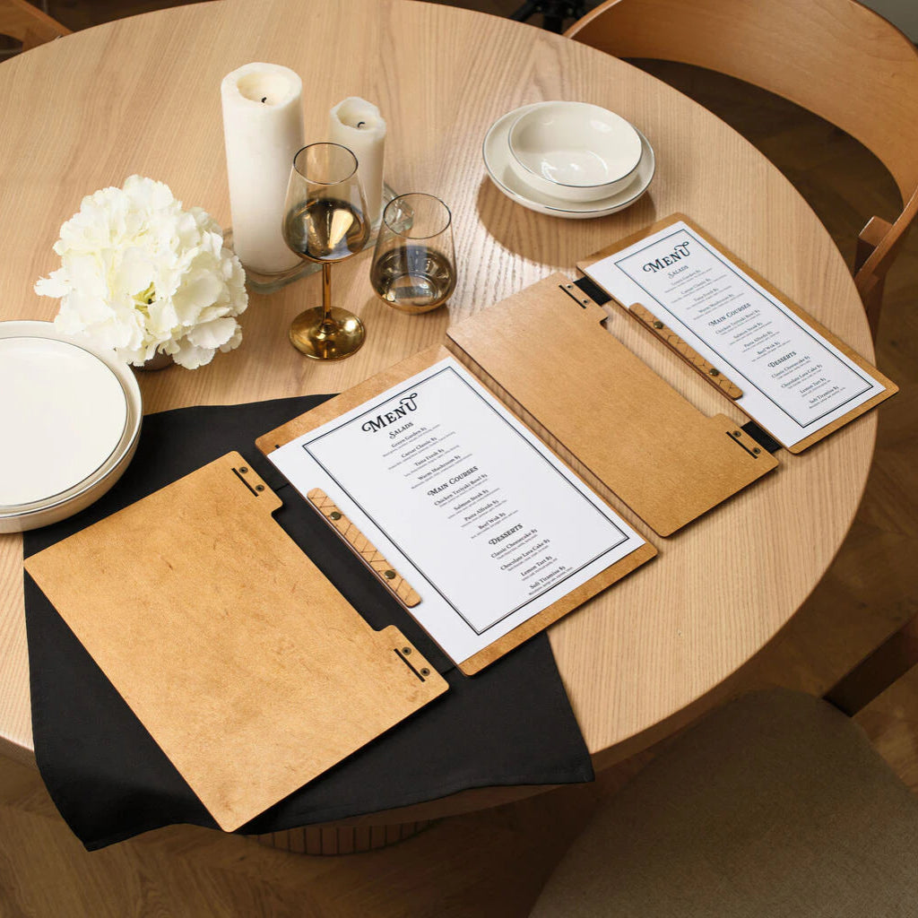 Two natural oak wooden menu boards on a set table, with one open to reveal a printed list of dishes.