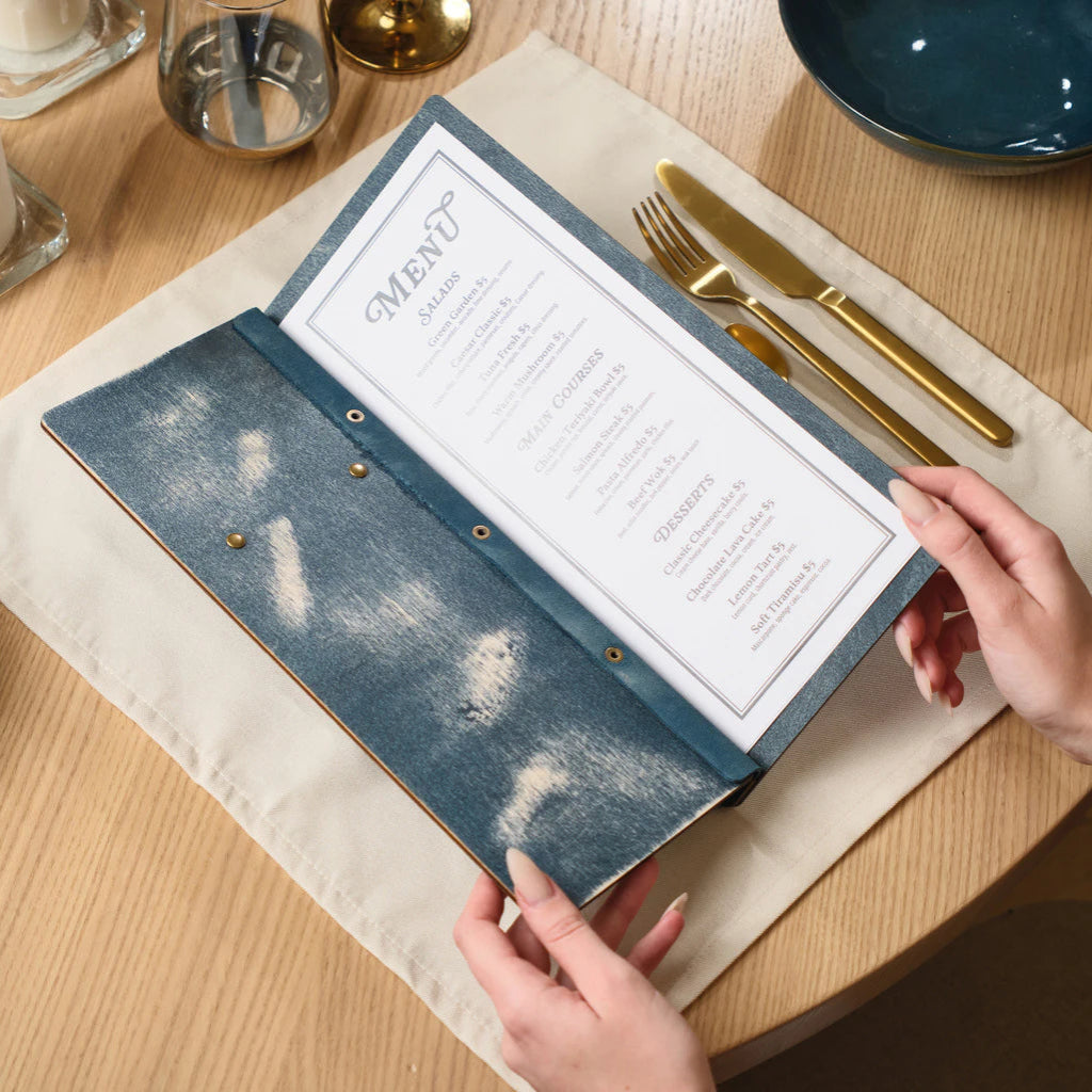A person holds open a wooden menu folder with a distressed "jeans" blue finish, revealing a printed paper menu with sections for Salads, Main Courses, and Desserts on a light wooden table set with gold cutlery.