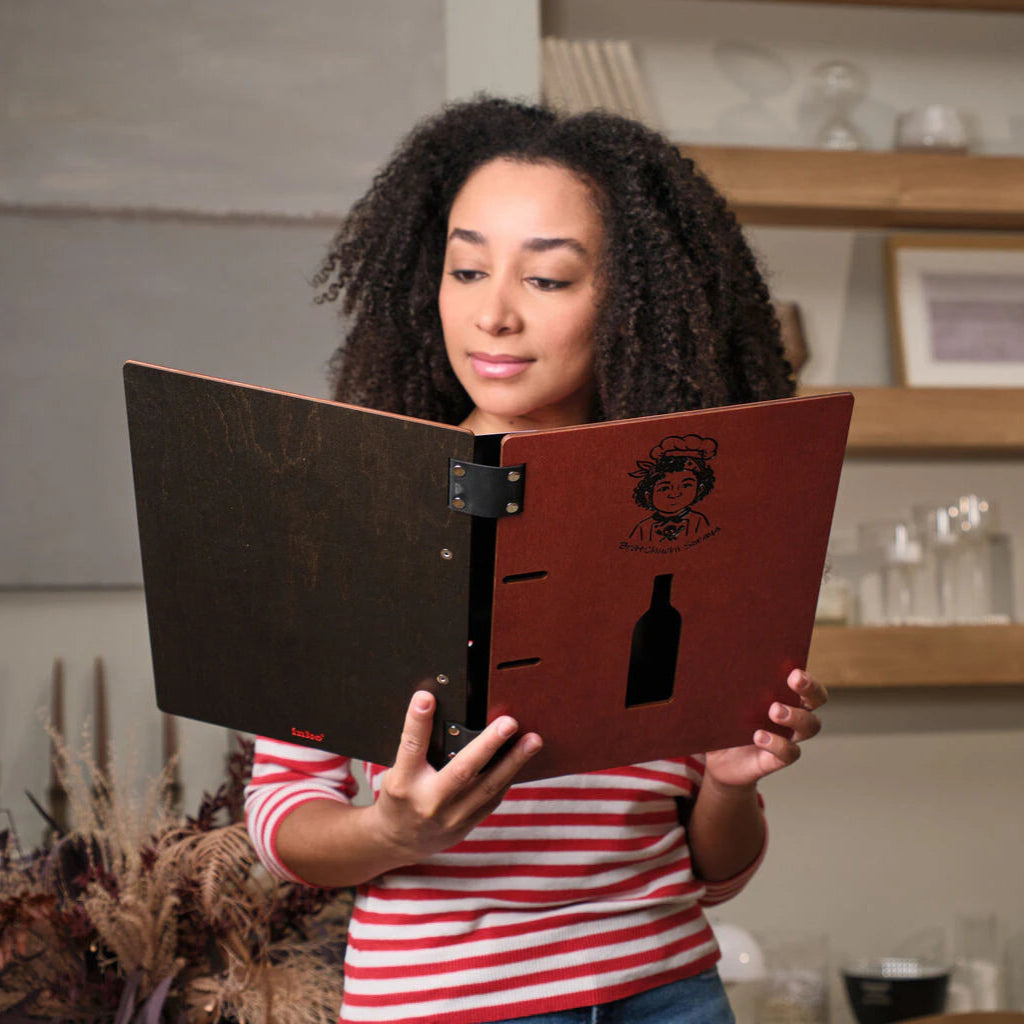 Frontal view of a woman holding an open chili-red wooden menu, highlighting the decorative engraving and the functional ring-binder design