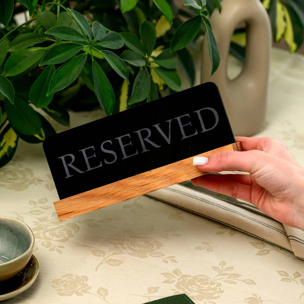 Modern black acrylic reserved sign with a natural wooden base held by a hand over a restaurant table setting
