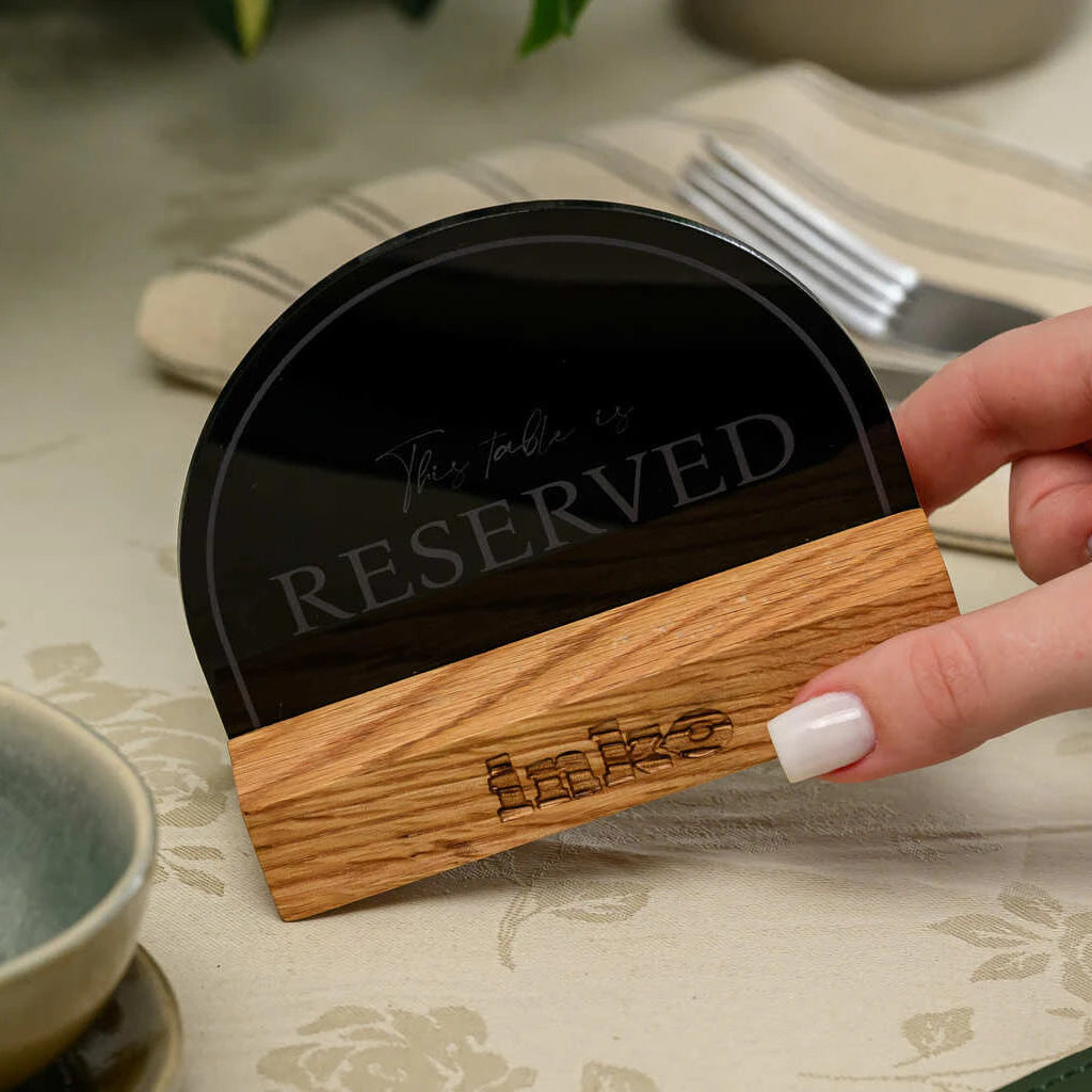 A close-up shot of a hand with white nails holding a 'Reserved' table sign at a slight angle. The sign consists of a black semicircular acrylic plate with the elegant engraving 'This table is RESERVED'. It is mounted on a light-colored oak wooden base featuring a small engraved logo