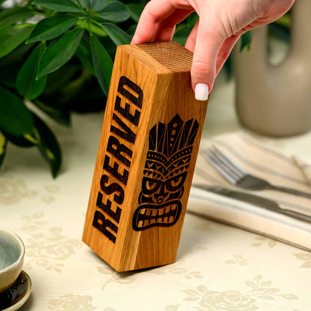A close-up of a person's hand placing a solid oak "Reserved" table sign. The vertical wooden block features a bold laser-engraved "RESERVED" text on one side and an intricate Tiki mask design on the other. The image highlights the natural wood grain and high-quality craftsmanship of this restaurant accessory