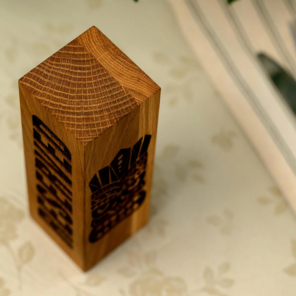 A high-angle, close-up shot of a solid oak "Reserved" table sign, highlighting the natural growth rings on the top cross-section. The vertical block features deep laser engraving of the word "RESERVED" and a Tiki mask on its sides. The warm, golden tones of the oak wood demonstrate the premium handcrafted quality of the product
