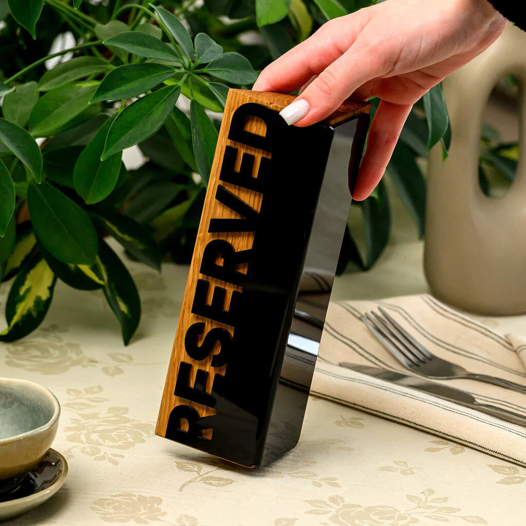 A hand with a white manicure holds a rectangular table sign vertically. The sign is made of light wood with a glossy black acrylic plate on top featuring the word 'RESERVED' in cutout letters. It stands on a floral tablecloth next to a striped napkin and silver cutlery