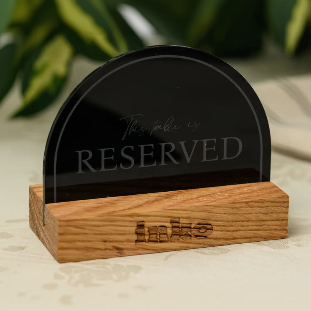 A tabletop reservation sign featuring a semicircular black acrylic plate held by a rectangular wooden block. The acrylic part has 'This table is RESERVED' engraved in an elegant font. The wooden base features a small engraved 'Inko' logo. It is placed on a light floral tablecloth.