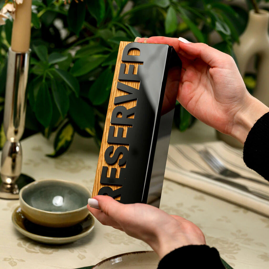 A person holds a rectangular 'Reserved' sign horizontally with both hands. The sign is made of oak-colored wood with a sleek black acrylic plate on top, featuring the word 'RESERVED' in bold cutout letters. It is shown against a lifestyle background with a set dining table and green houseplants