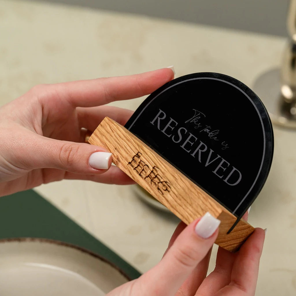 A close-up shot of a person's hands holding a modern restaurant reservation sign. The sign features a semicircular black acrylic plate with the engraved text 'This table is RESERVED' in an elegant font. The plate is mounted on a light-colored wooden base that has a small logo engraved on the side.