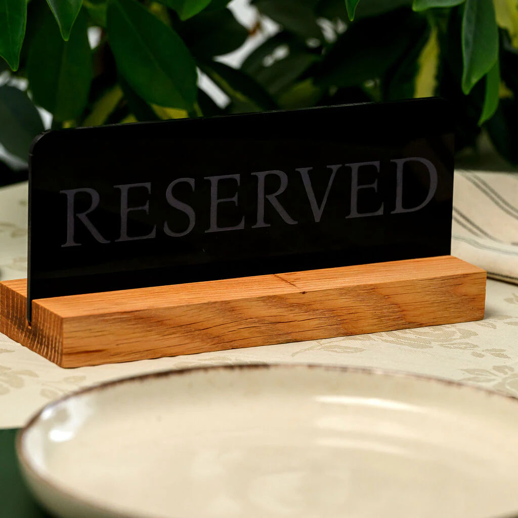 Elegant black acrylic "Reserved" sign with a natural oak wood base standing on a restaurant table next to a ceramic plate