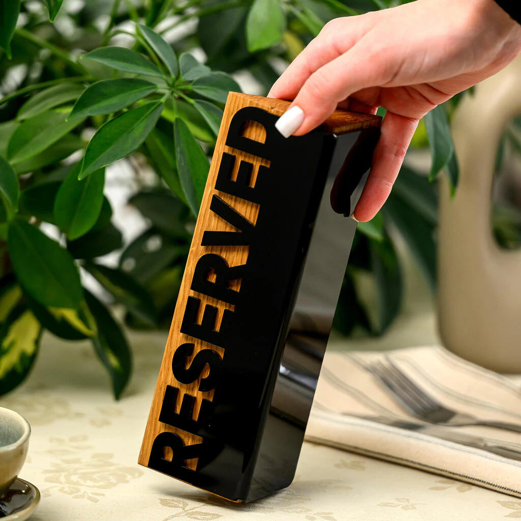 A close-up shot of a hand with a white manicure steadying a vertical 'Reserved' sign. The sign is made of light-colored wood with a glossy black acrylic plate where the word 'RESERVED' is cut out. It sits on a floral tablecloth next to a striped napkin with cutlery and green plants in the background.