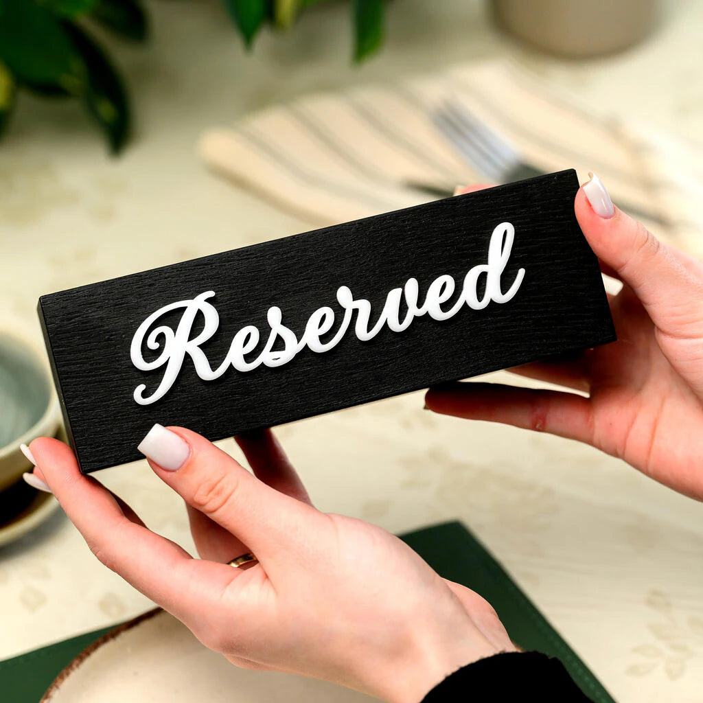 A horizontal "Reserved" table sign held in two hands to demonstrate its scale. The sign is made of high-quality black-stained wood with a prominent wood grain texture and features elegant 3D white acrylic script lettering. It is displayed in a restaurant setting with a neutral tablecloth and greenery in the background