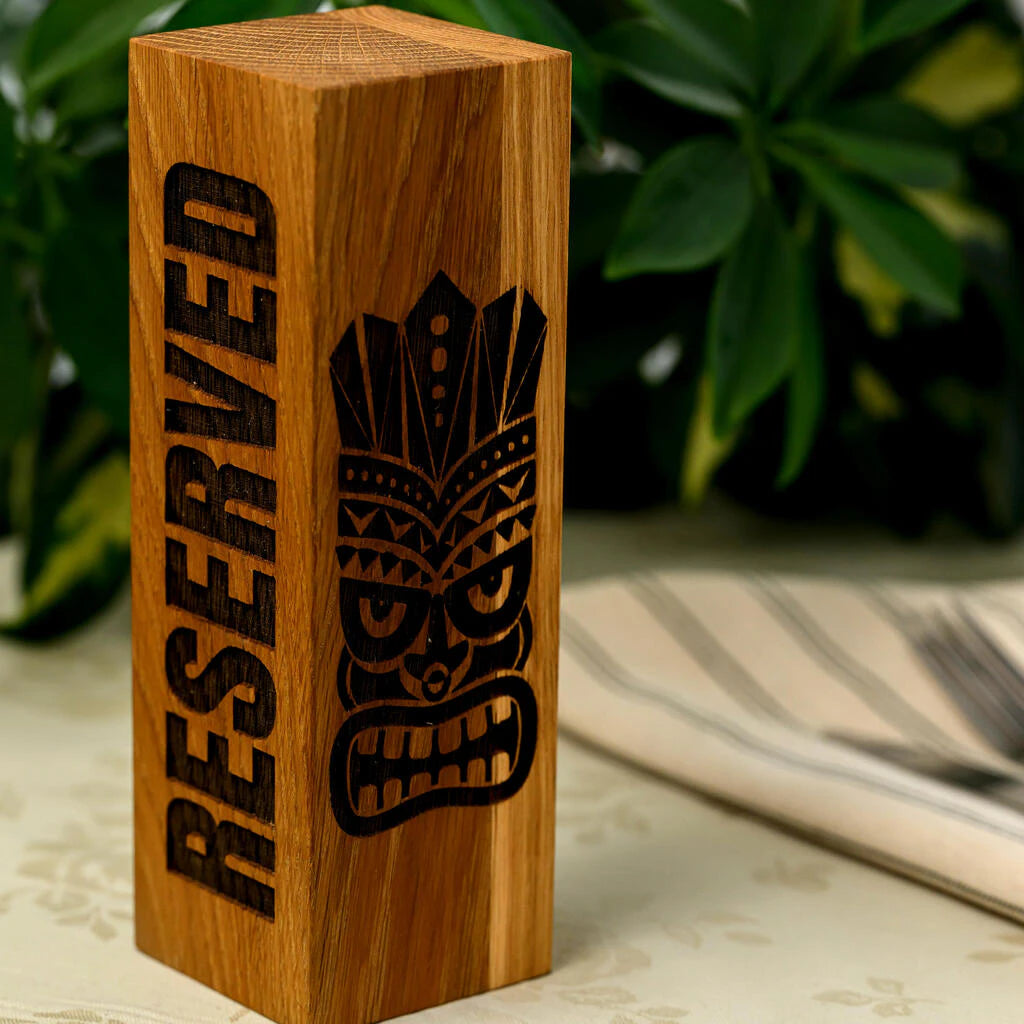 A tall, rectangular "Reserved" table sign handcrafted from solid oak wood. One side features the word "RESERVED" in a bold, vertical engraved font, while the adjacent side showcases a detailed, laser-engraved Tiki mask. The sign stands on a restaurant table with a tropical plant in the background