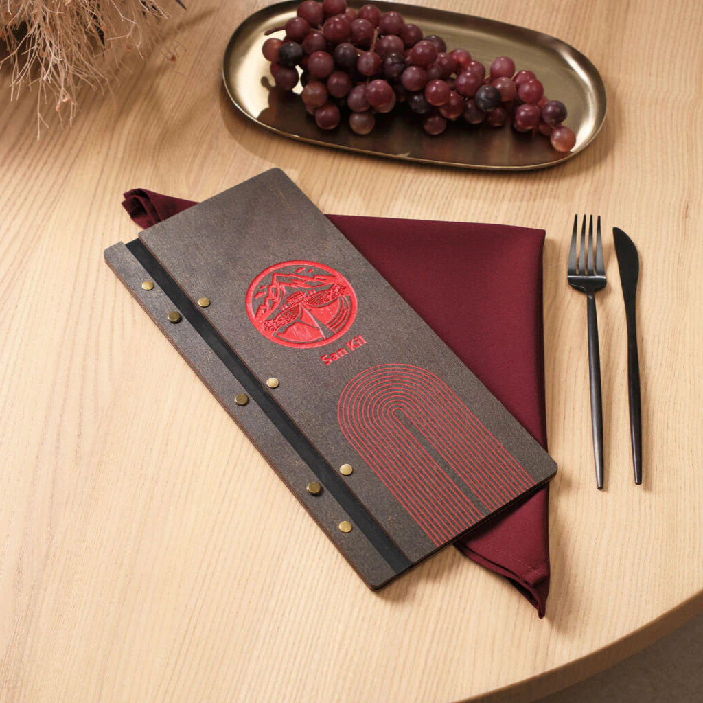 A wenge-colored wooden menu folder lying on a burgundy napkin, featuring a vibrant red circular logo with mountain motifs, the "San Kil" brand name, and a stylized arch pattern. The dark wood finish is complemented by antique brass rivets and a black leather spine.
