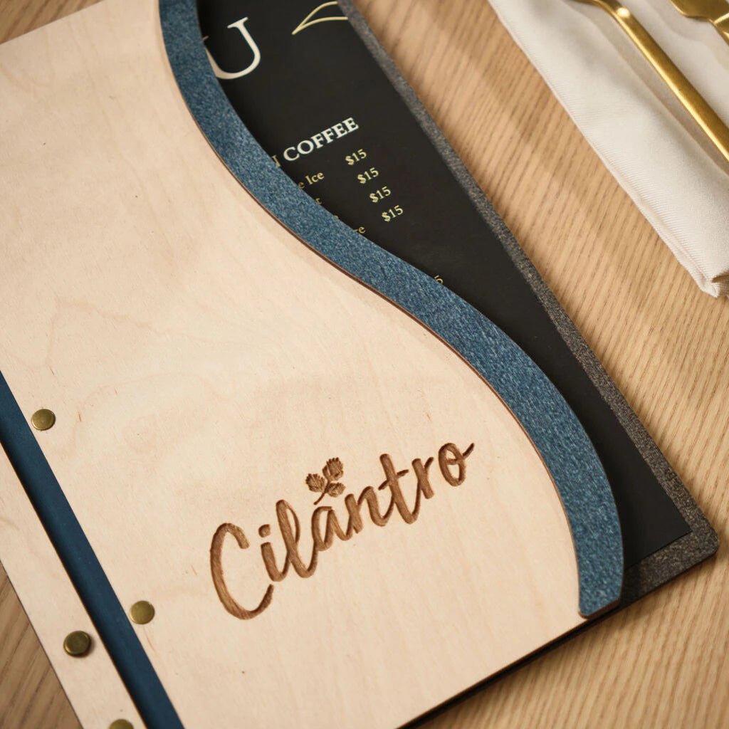 Close-up of the "Cilantro" laser-engraved logo on a handcrafted Ivory wooden menu cover with professional brass screw posts and a blue wavy border