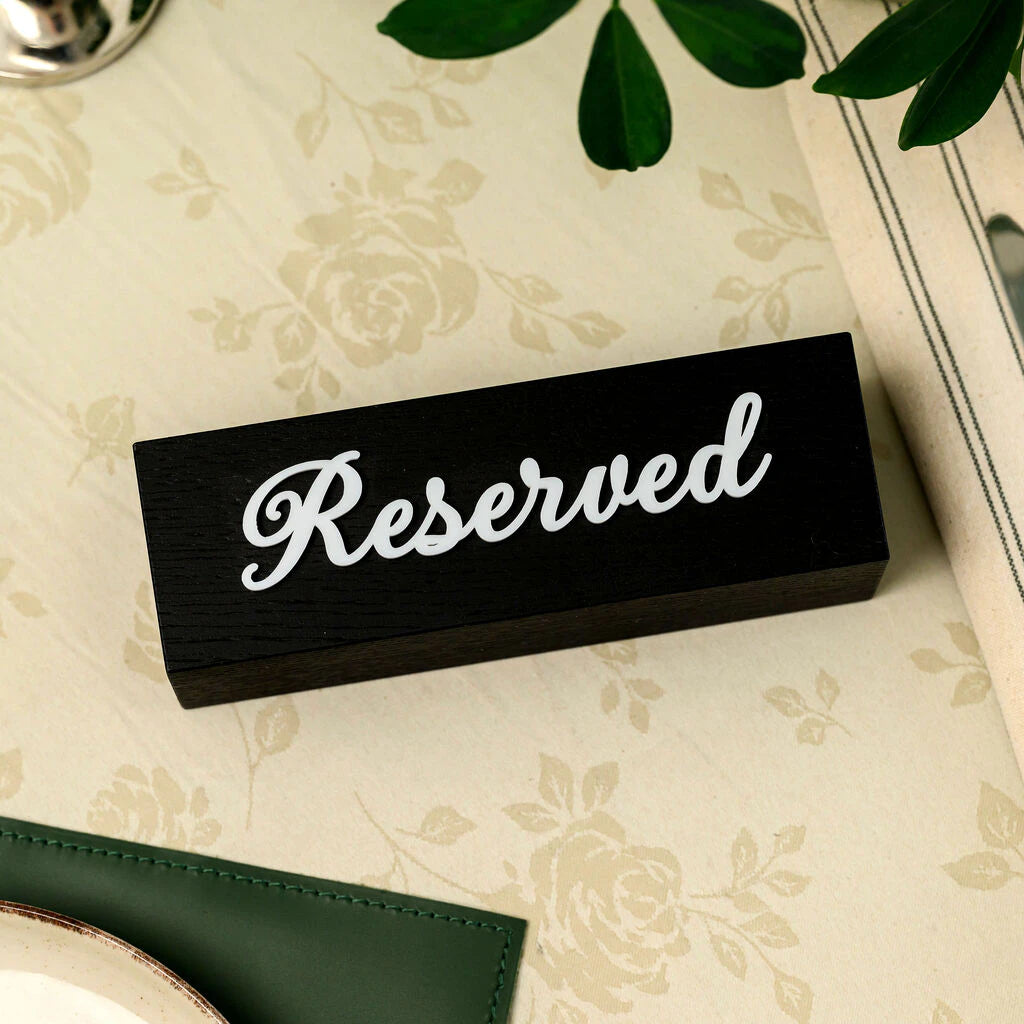 A high-angle, close-up photograph of a professional wooden "Reserved" table sign. The sign is made of high-quality black-stained wood with visible wood grain texture. It features elegant, applied 3D white acrylic script lettering for the word "Reserved". It rests on a beige vintage-style cloth with a subtle leaf pattern, surrounded by blurred table items like cutlery and plants
