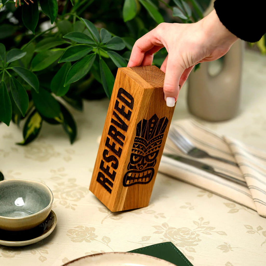 Handcrafted oak "Reserved" table sign with Tiki mask engraving, held by a person in a restaurant setting