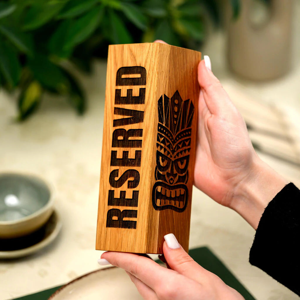 A close-up photograph showing a person's hands with a white manicure holding a handcrafted solid oak wood table sign. The vertical sign features deep laser engraving: the word "RESERVED" on one face and a detailed Tiki mask design on the other. It's set in a tropical cafe environment with lush green plants in the background