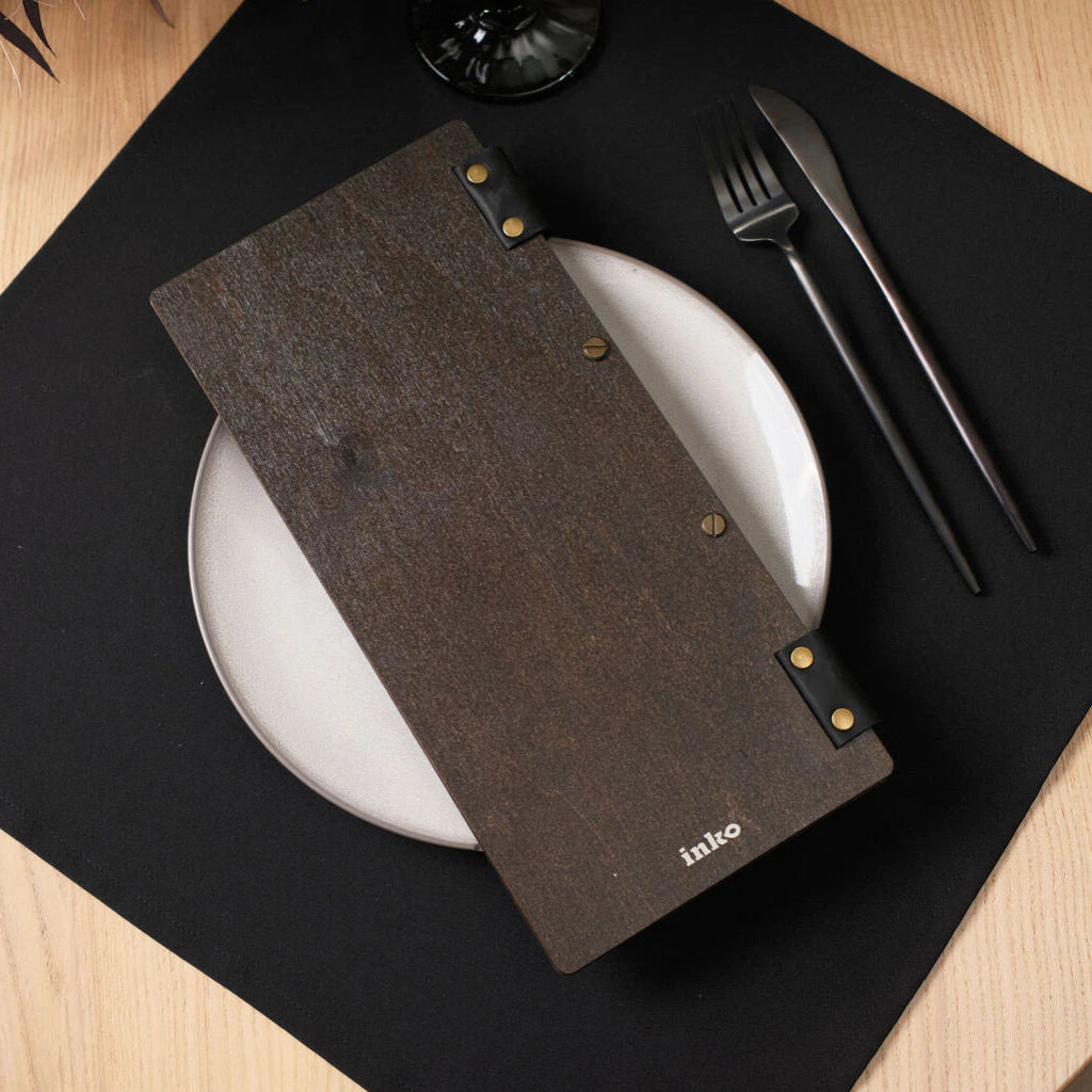 The back of the wenge-colored wooden menu folder is shown resting on a white plate, featuring brass binding screws, black leather accents with rivets, and a small white "inko" logo at the bottom