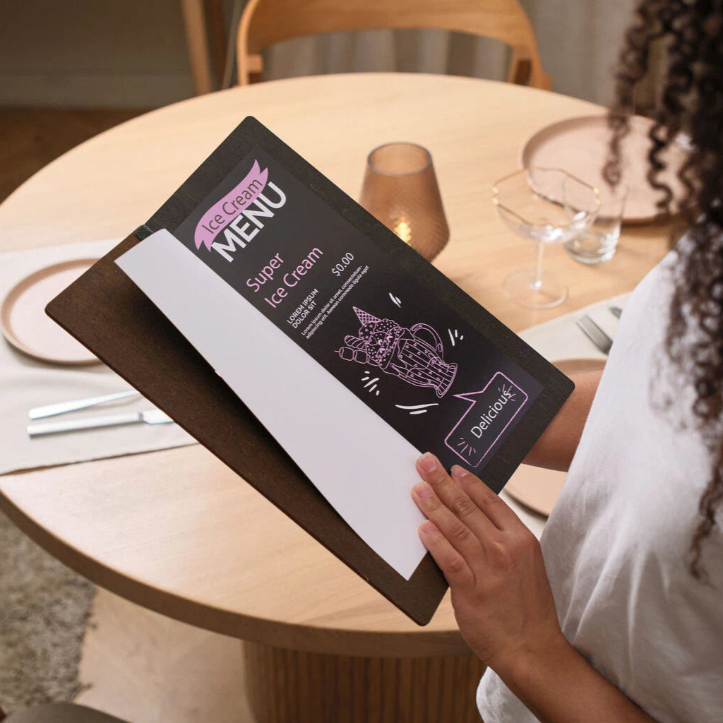 A person holds a dark wooden menu board over a set table, displaying a black "Ice Cream Menu" insert with white and pink text and a stylized illustration of a milkshake.
