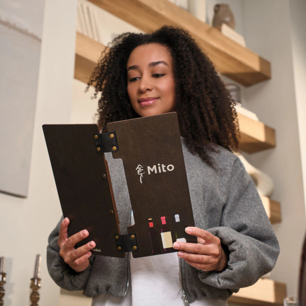 A woman with curly hair holds open a dark wenge wooden menu folder featuring a white "Mito" logo and colorful UV-printed wine bottle illustrations. The folder is bound with black leather straps and brass rivets.