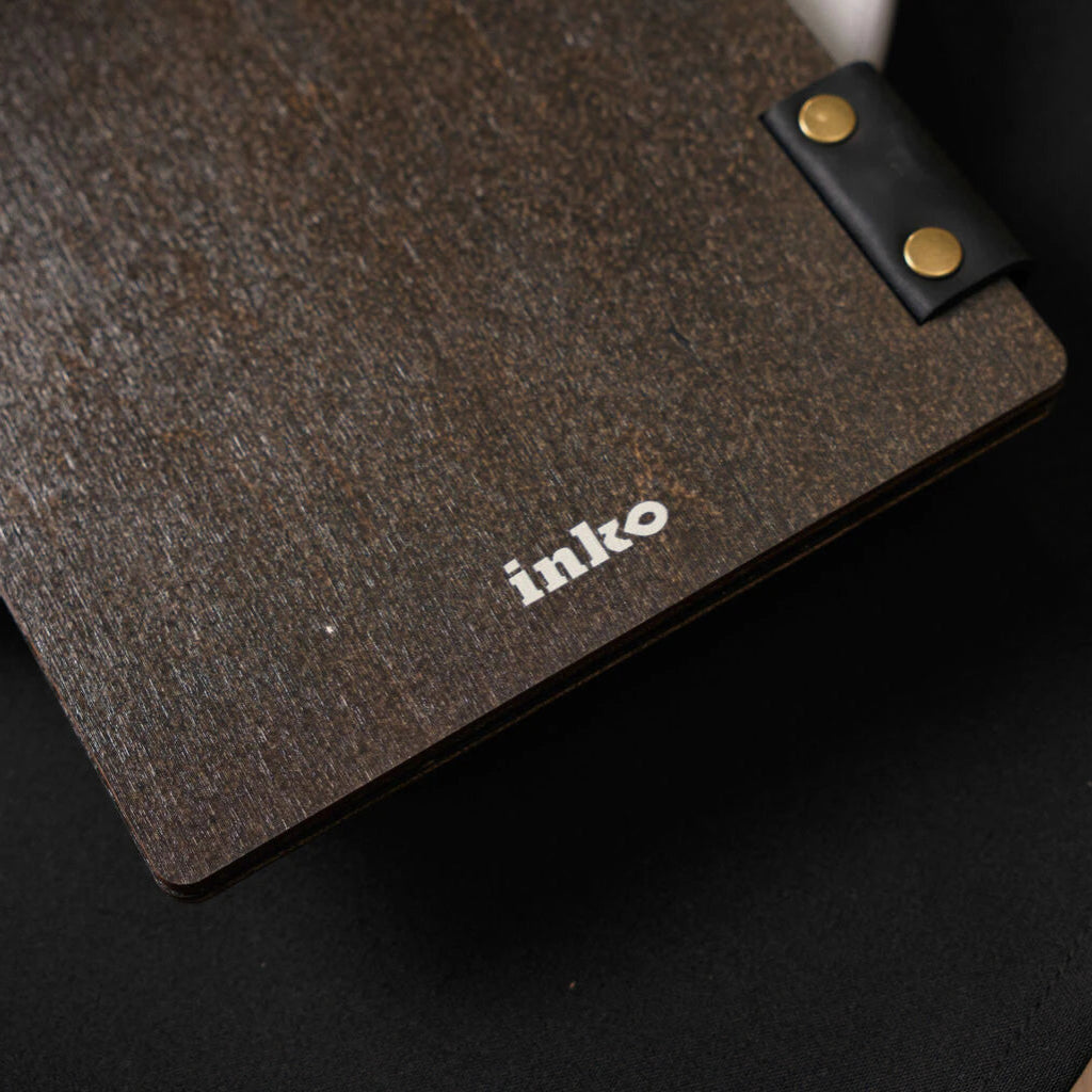 A close-up shot of the back of a dark wenge wooden menu folder, showing the textured wood grain and a small white "inko" logo UV-printed at the bottom. A black leather strap with brass rivets is visible at the edge.