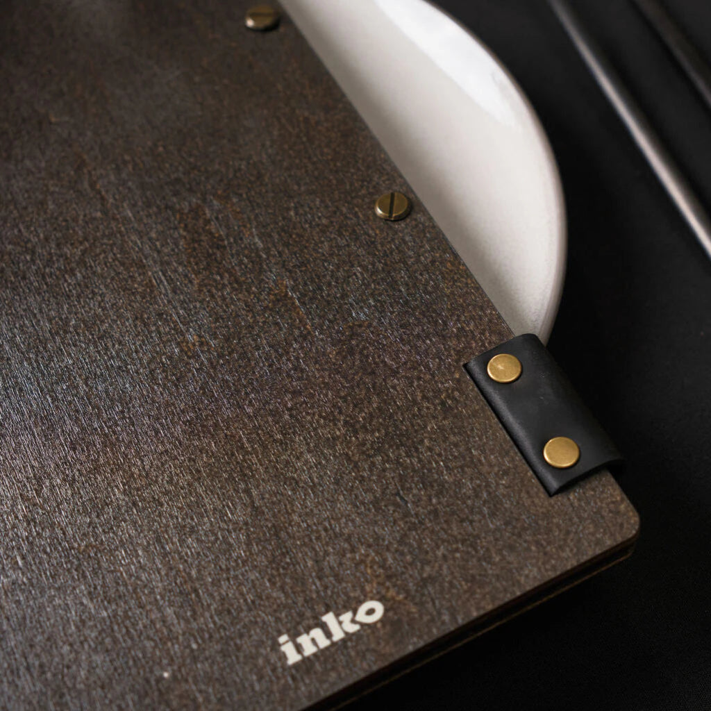 A close-up view of a dark wenge wooden menu's edge, showing the "inko" logo and the black leather binding strap with two gold-toned rivets. Metal binding screws are visible along the spine.