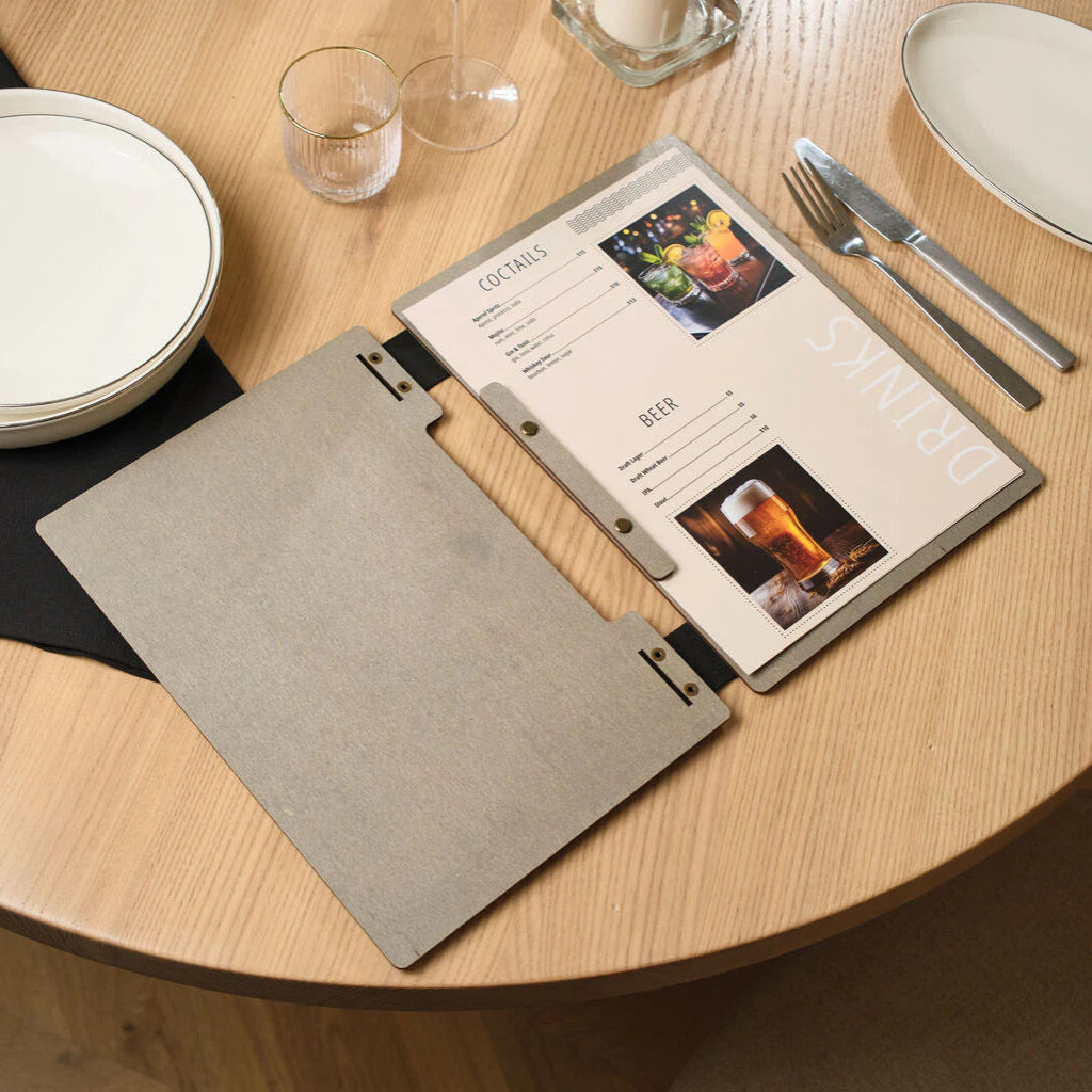 An open grey wooden menu folder on a wooden table, showing a "COCKTAILS" and "BEER" drinks list with full-color food and beverage photography.