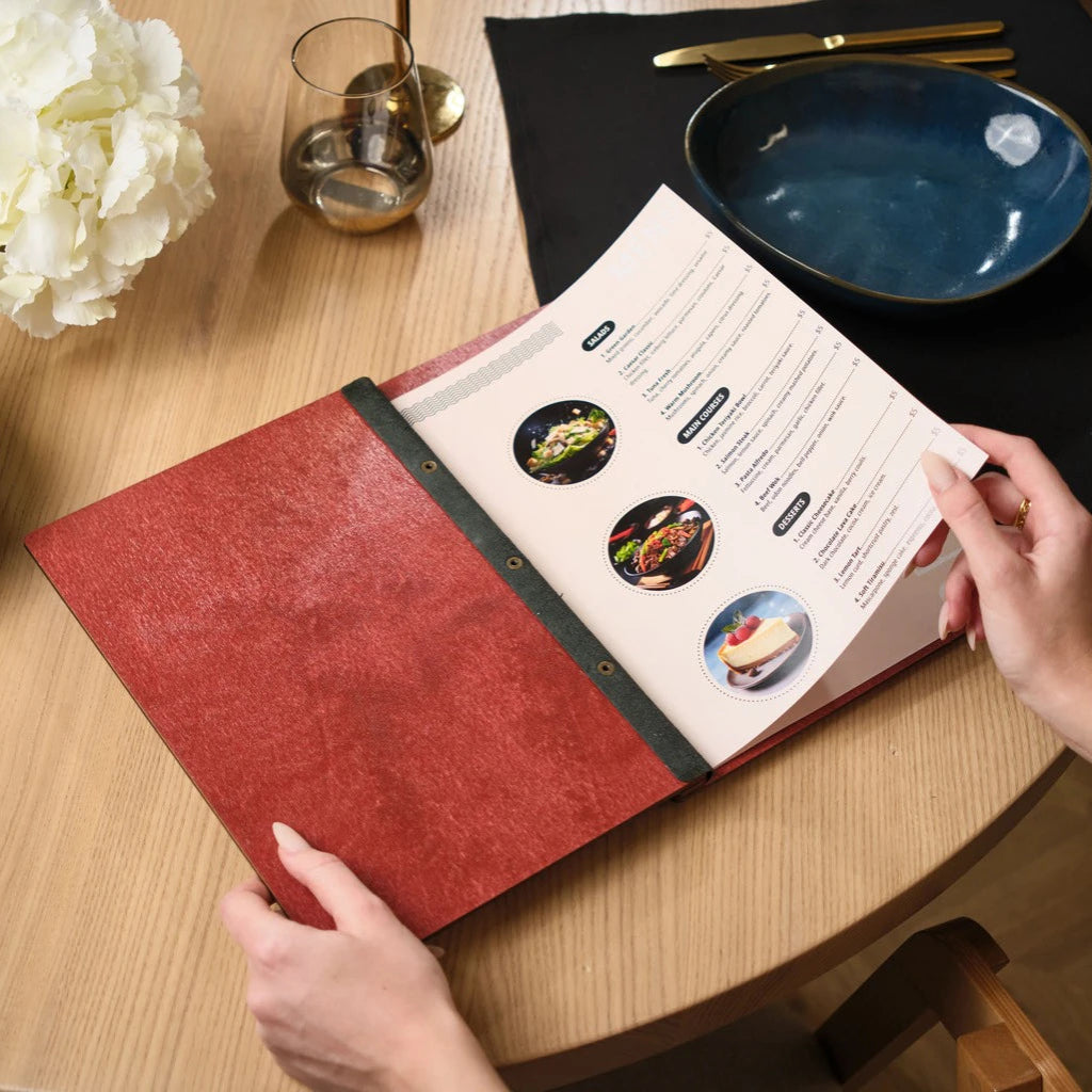 Internal view of an open red wooden menu folder held by a customer, showcasing printed menu inserts for a restaurant. The handcrafted wood menu cover features a secure black leather hinge and professional bronze bolt binding