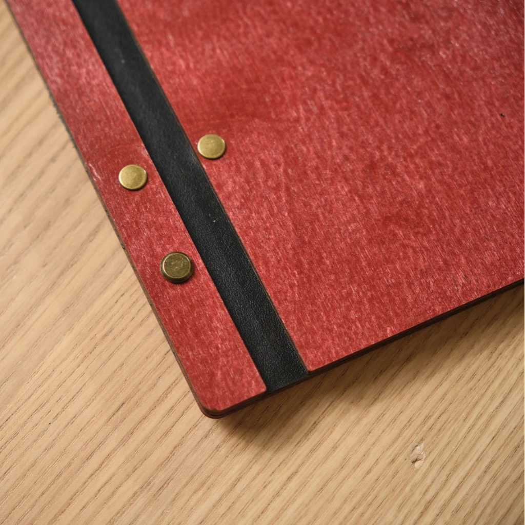Detail view of a handcrafted wood menu cover in vibrant red with a flexible black leather spine. The design is secured with decorative gold-toned metal fasteners, showcasing the high-quality finish and materials