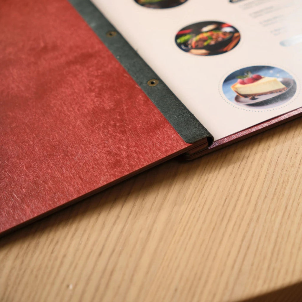 A detailed macro shot of a premium red wood menu holder, focusing on the durable leather spine and gold-toned metal fasteners. The image showcases the high-quality craftsmanship and elegant wood grain texture on a modern dining table
